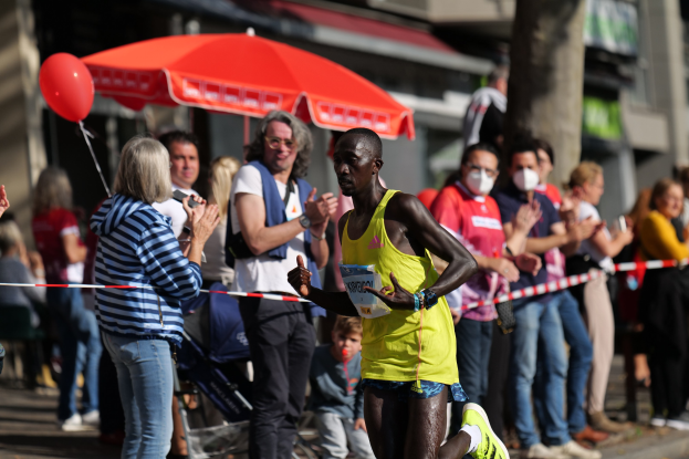 Ein Mann in einem gelben T-Shirt und Laufschuhen sprintet durch einen Marathon, während ihn Zuschauer umringen und klatschen, mit einem Schirm und einem Kinderwagen im Hintergrund.