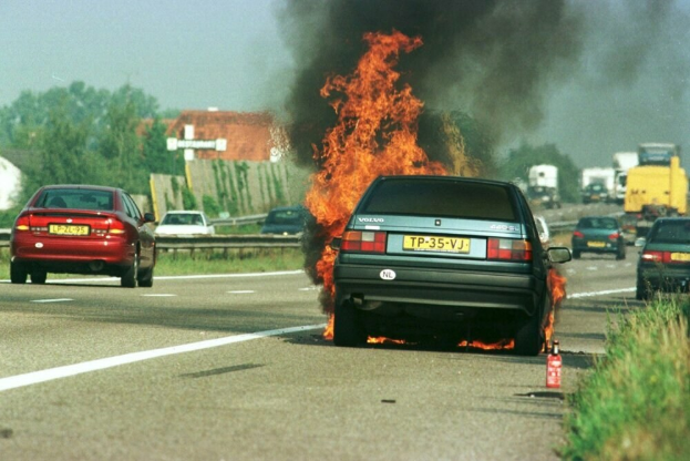 Ein Auto, das in Flammen steht, am Straßenrand umgeben von anderen Fahrzeugen, mit Bäumen, Gebäuden und einem klaren blauen Himmel im Hintergrund und Gras mit einem Feuerlöscher auf der rechten Seite.