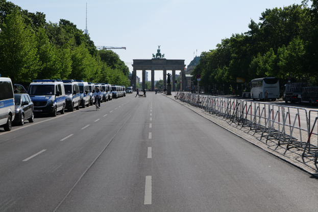 Eine Reihe von Polizeiwagen auf einer Straße vor dem Brandenburger Tor in Berlin geparkt, mit Menschen auf Fahrrädern und Stehenden, Barrieren, Bäumen und einem Bogen mit Statuen im Hintergrund.