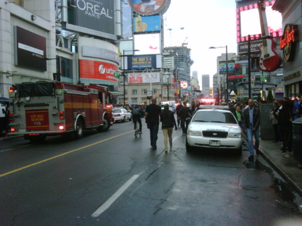 Eine belebte Straße mit parkenden Fahrzeugen auf beiden Seiten, Menschen, die auf der Straße und dem Gehweg gehen, und Geschäfte auf beiden Seiten.
