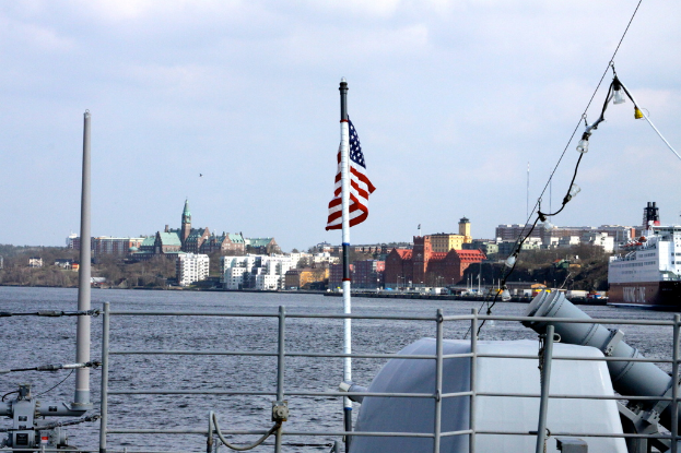 Eine Stadtansicht mit einem Schiff im Vordergrund, das eine Flagge zeigt, Booten auf dem Wasser, Gebäuden, Bäumen und Pfählen im Hintergrund, unter einem bewölkten Himmel, mit Wasser unten.