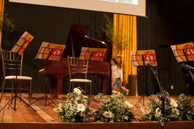 Eine Bühne mit Mikrofonen auf Ständern, Blumensträußen, Stühlen, Notenpulten mit Büchern, einem Flügel, einer Person, die läuft, und einem Hintergrund mit Bäumen, Vorhängen und einer Projektionswand.