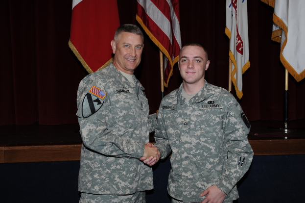 Zwei Personen in Uniformen geben sich die Hand und lächeln, während sie vor vier Fahnen auf Stangen vor einem dunklen Hintergrund stehen.