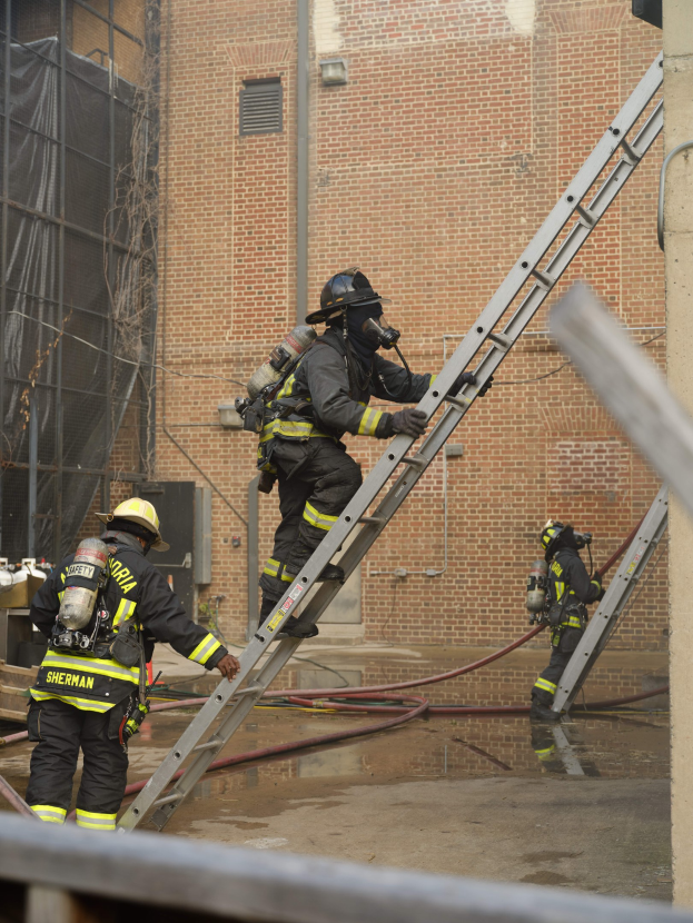 Feuerwehrleute in Helmen und Ausrüstung klettern an einer Leiter vor einem Backsteingebäude mit Rohren auf dem Boden und einer Metallstange am unteren Ende, mit einem weiteren Gebäude mit Fenstern und einem Netz im Hintergrund.