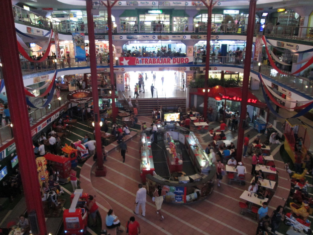 Ein belebter Einkaufszentrum-Innenraum mit Säulen, Menschen, Tischen, Fliesen, Flaggen, Geschäften, einem Banner, Treppen und einem Namensschild.