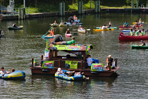 Eine Gruppe von Menschen in kleinen Booten auf dem Wasser, einige halten Paddel, mit Schildern und verschiedenen Gegenständen im Hintergrund.
