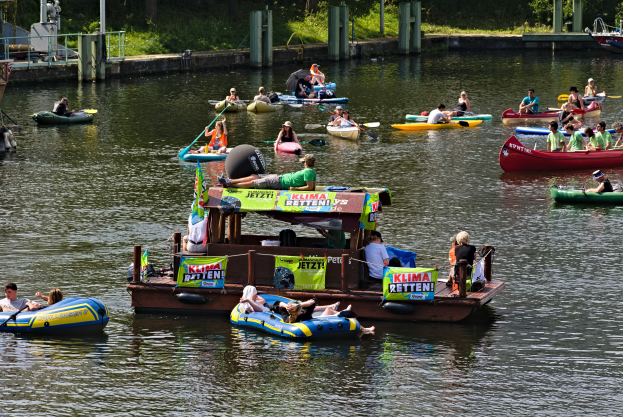 Eine Gruppe von Menschen in kleinen Booten auf dem Wasser, einige halten Paddel, mit Schildern und verschiedenen Gegenständen im Hintergrund.