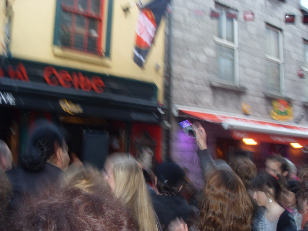 Ein unscharfer Bild mit Menschen auf der Straße unten und Gebäuden, Lichtern, Fenstern und einer Flagge oben.