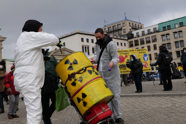 Eine Gruppe von Menschen in weißen Schutzanzügen und Masken steht um einen gelben Eimer herum, mit einem Wagen im Vordergrund, hält Schilder während einer Anti-Atom-Protest in Berlin, mit Gebäuden, Fahnen und einem bewölkten Himmel im Hintergrund.