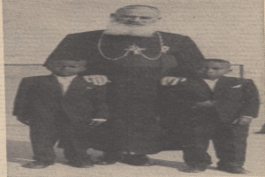 Ein Schwarz-Weiß-Foto eines Mannes, der neben zwei Jungen in Blazern und Krawatten steht, mit einem Text unten, der darauf hinweist, dass er ihr Vater ist.