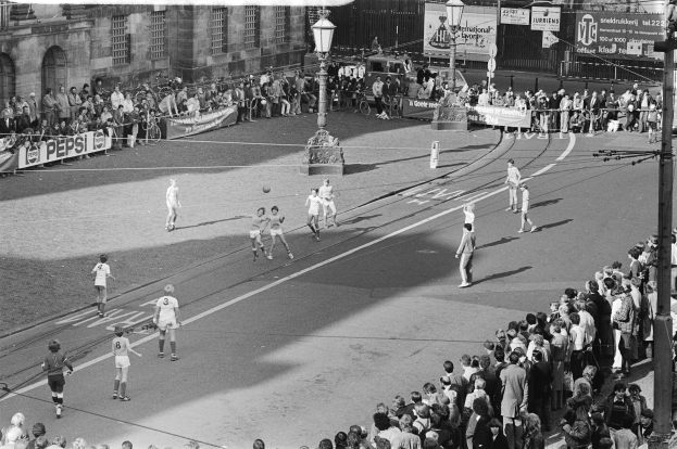 Schwarzes Foto eines auf der Straße gespielten Fußballspiels mit Zuschauern, umgeben von Laternenmasten, Gebäuden und Bannern.