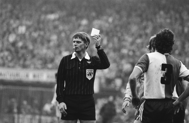 Schwarzes und weißes Bild eines Schiedsrichters, der eine rote Karte in der Luft hält, vor einer Menge von Menschen in Fußballuniformen, mit einem Zaun im Hintergrund.