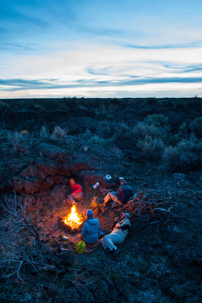 Ein Lagerfeuer mit Menschen, die darum sitzen und liegen, mit Mützen tragend, einem Beutel und anderen Gegenständen auf dem Boden, umgeben von Bäumen und Himmel.
