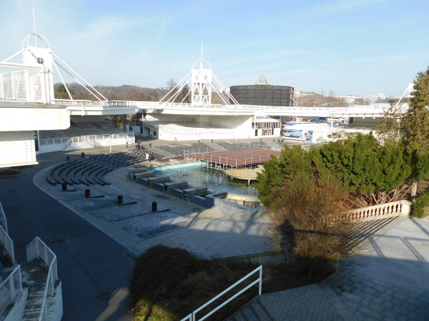Blick auf das Olympische Stadion von einem hohen Standpunkt aus, mit verschiedenen Objekten und Grün im Vordergrund und einem bewölkten Himmel im Hintergrund.