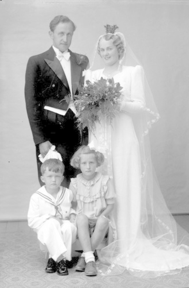 Schwarzes und weißes Foto einer Braut in einem weißen Kleid mit einem Strauß, die eine Braut hält, ein grinsender Bräutigam neben ihr und zwei Kinder, die vor ihnen auf dem Boden sitzen.