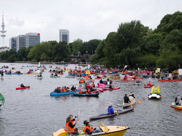 Eine große Gruppe von Menschen in Kanus auf einem Gewässer, einige halten Paddel und Fahnen, im Hintergrund sind Bäume, Gebäude und ein Turm zu sehen.