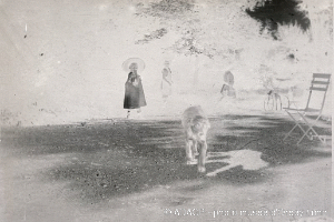 Ein altes Schwarz-Weiß-Foto einer Frau, die ihren Hund in einem Park spazieren führt, mit Menschen, einem Fahrrad, einem Stuhl und Bäumen im Hintergrund und Text am unteren Rand.