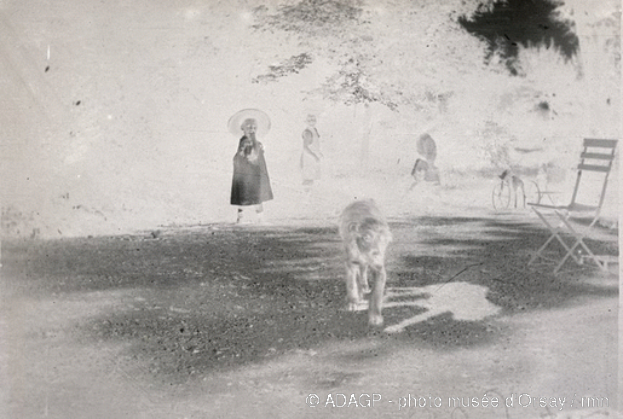Ein altes Schwarz-Weiß-Foto einer Frau, die ihren Hund in einem Park spazieren führt, mit Menschen, einem Fahrrad, einem Stuhl und Bäumen im Hintergrund und Text am unteren Rand.