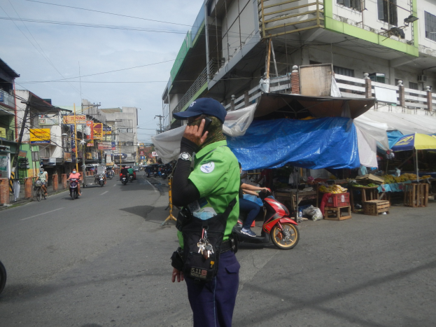 Ein Polizeibeamter in grüner und blauer Uniform mit blauer Mütze steht auf einer Straße und spricht in ein Handy, mit Fahrzeugen und Marktständen im Hintergrund und einem Gebäude im bewölkten Himmel.