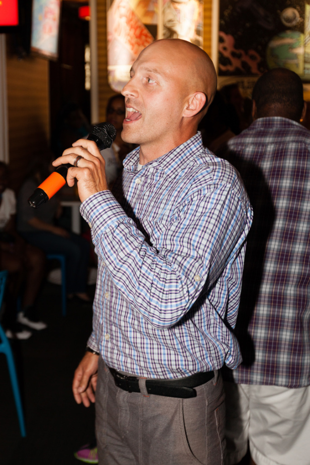 Eine Person in einem schwarzen Gürtel steht und singt in ein Mikrofon mit einer Gruppe von Menschen hinter ihr.