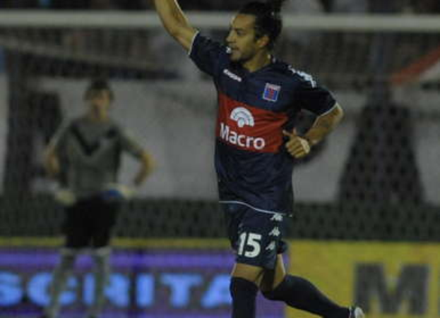 Ein Fussballer feiert nach einem Tor auf dem Boden, mit einer jubelnden Menge und einer Person im Hintergrund, alles vor einem unscharfen, energetischen Hintergrund.