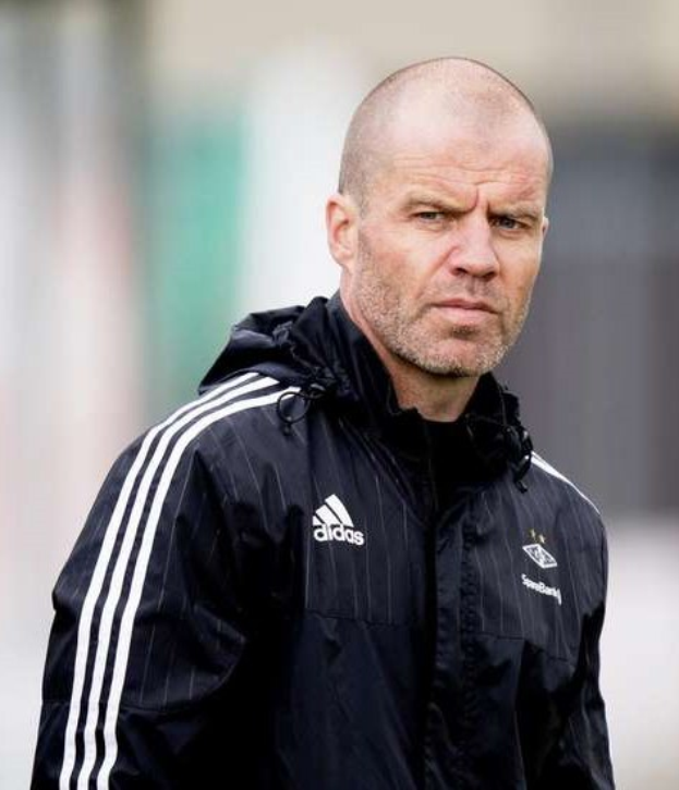 Baldiger Trainer in einer blauen Jacke mit weißen Streifen und einem weißen Kragen, der auf einem Fußballfeld steht und die Hände gefaltet hat.