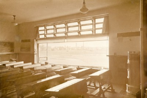 Schwarzer und weißer Klassenraum mit hölzernen Kirchenbänken, Tischen, einem Kühlschrank, einer Wandtafel, einem Vorhangfenster und Deckenleuchten.