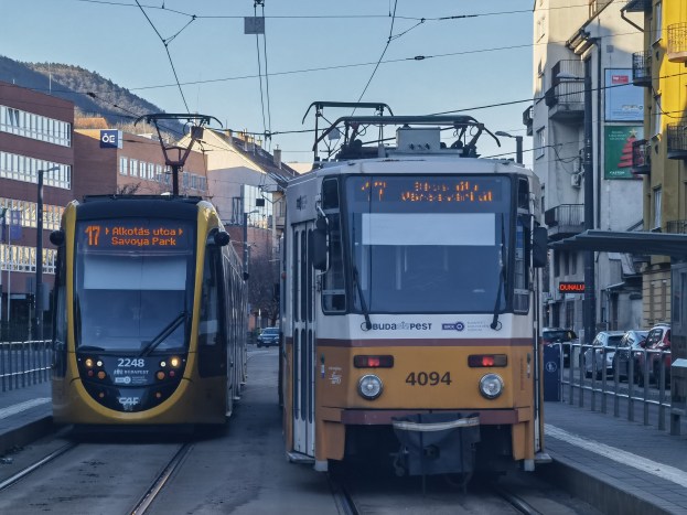 Zwei gelbe und weiße Straßenbahnen auf einer Stadtstraße mit hohen Gebäuden, Fahrzeugen, Geländern, Bäumen und einem Hügel im Hintergrund.