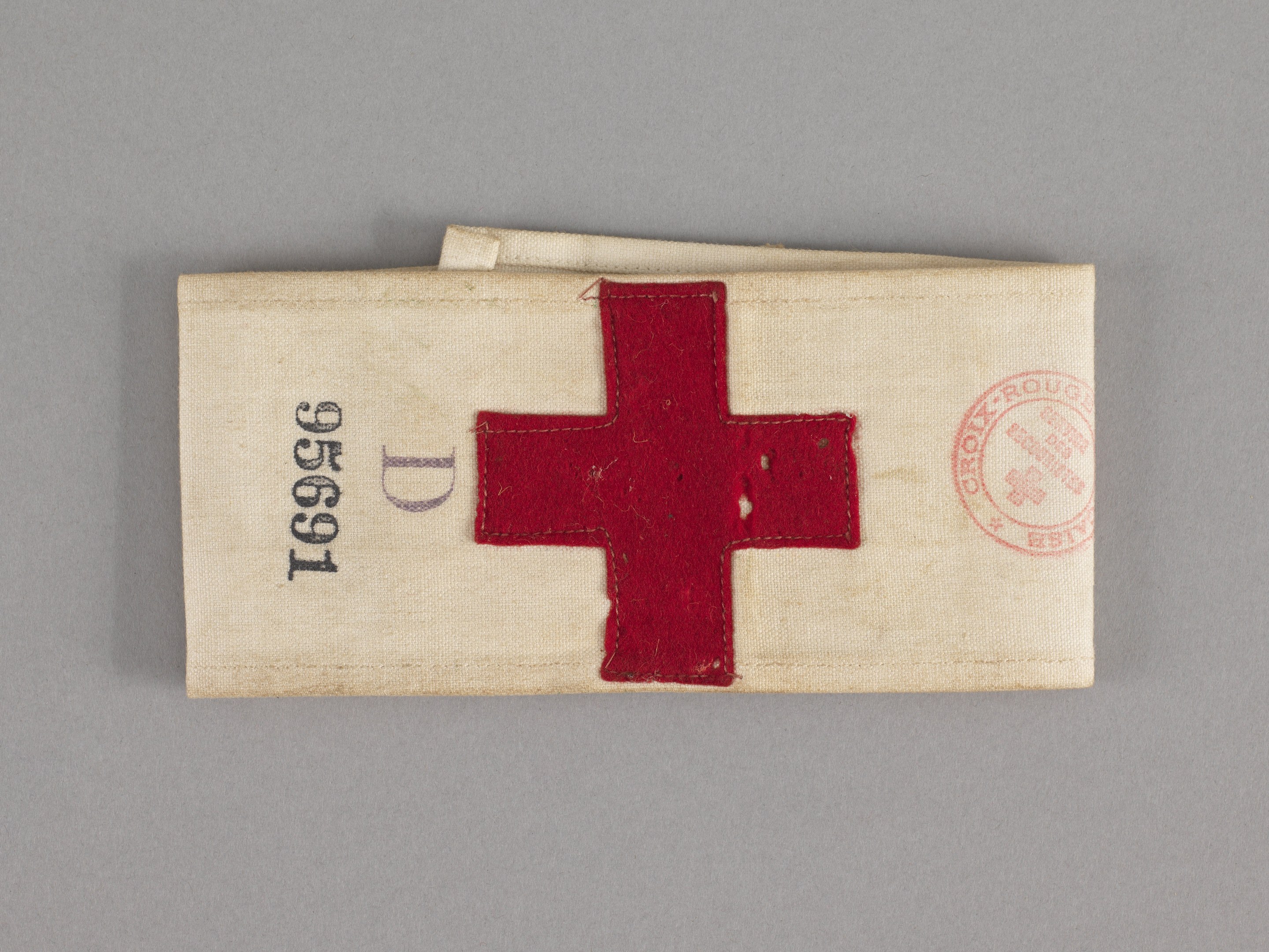 Ein rotes Kreuz-Ersthilfepaket in einem Stoffbeutel mit Text und einem Stempel, das auf einer Oberfläche liegt.