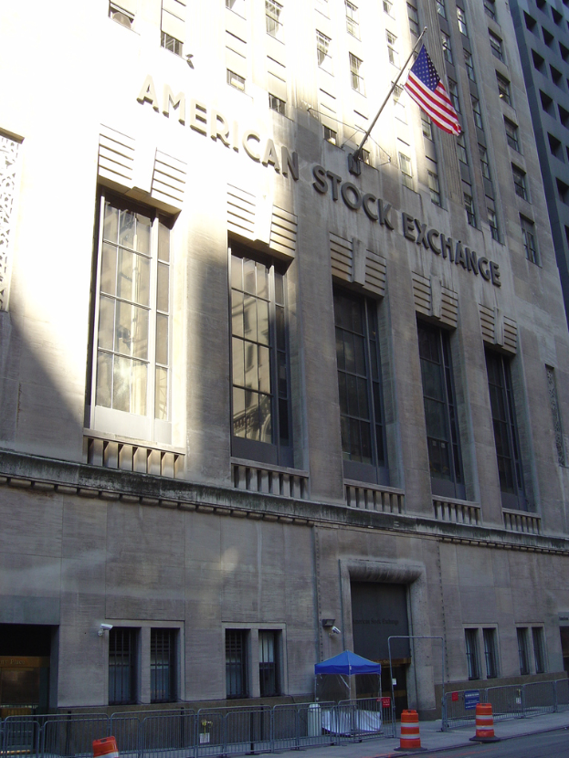 Ein Gebäude mit Text auf der Fassade, eine Fahnenstange an der Wand, ein Geländer auf dem Gehweg davor, Barrieren jenseits des Geländers und eine Straße im rechten unteren Bereich.