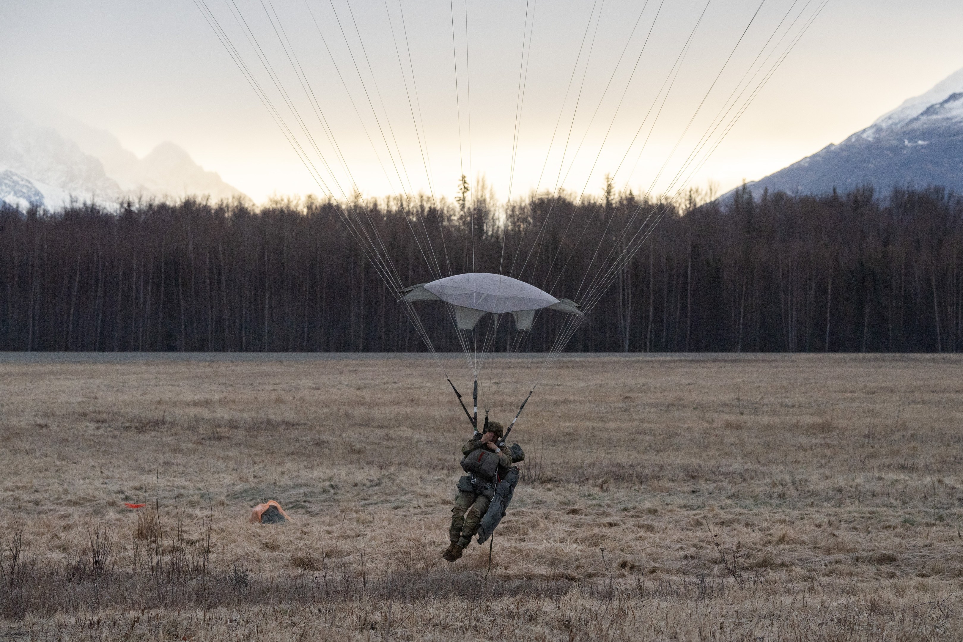Eine Person Paragleiten über einer grünen Wiese mit schneebedeckten Bergen im Hintergrund, die einen Helm trägt und von Bäumen umgeben ist unter einem klaren blauen Himmel.