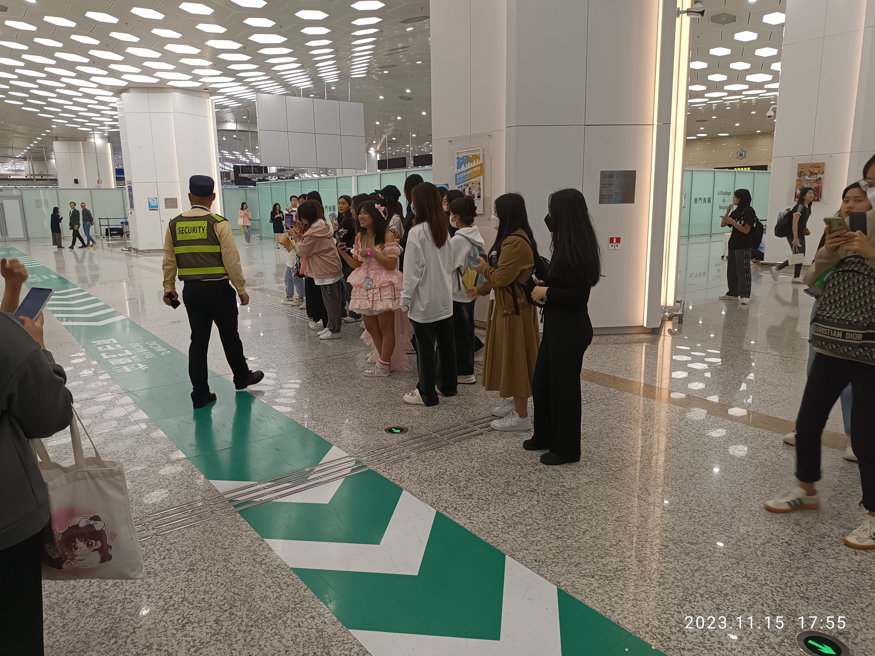 Menschen in der Schlange an einem Flughafen-Sicherheitscheck, mit Taschen und Mobiltelefonen, im Hintergrund Säulen und Deckenleuchten.