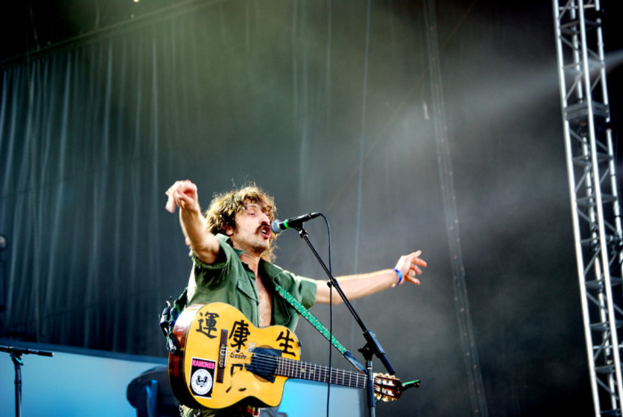 Ein Mann singt in ein Mikrofon, während er eine Gitarre hält und seine Hände ausbreitet, vor einem schwarzen Vorhang mit einer Eisenstange.
