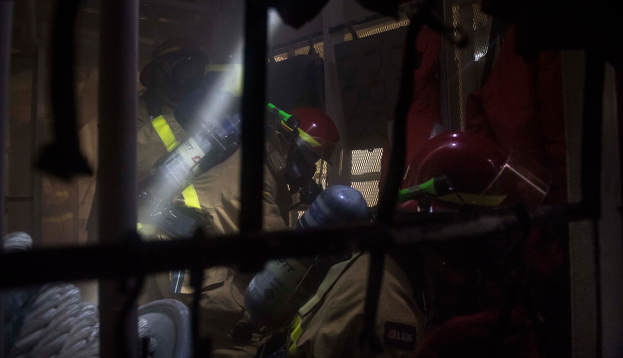 Eine Gruppe von Feuerwehrleuten in Helmen und Schutzausr√ľstung, einer mit einem Feuerl√∂scher, mit einem Metallgitter und einem Seil im Vordergrund und verschiedenen Gegenst√§nden im Hintergrund.