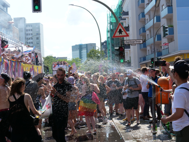 Menschen bei einer Gay-Pride-Veranstaltung, die sich gegenseitig mit Wasser bespritzen, während sie Gegenstände halten, mit einem Banner links und Gebäuden, Bäumen und Ampeln im Hintergrund.