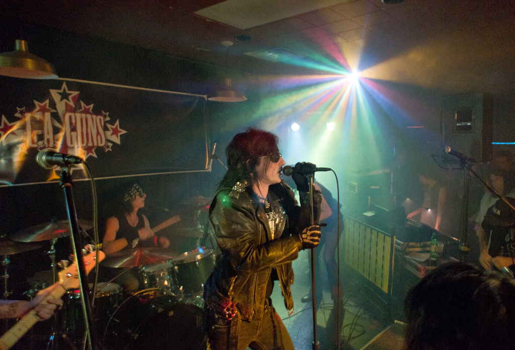 Eine Frau in einer schwarzen Jacke singt in ein Mikrofon auf der Bühne, mit einem Mann in Sonnenbrille und Stirnbinde, der ein Instrument spielt, und einem Plakat mit der Aufschrift "LA Guns" im Hintergrund, unter Diskoleuchten und Hängelampen.