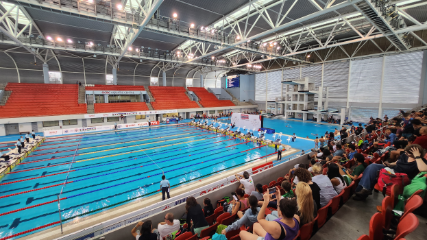 Ein überfülltes Schwimmbad mit Menschen auf Stühlen und einigen im Wasser Stehenden, umgeben von Tafeln mit Text, unter einer Decke mit Lampen und Eisenstangen im Olympischen Schwimmzentrum in London.
