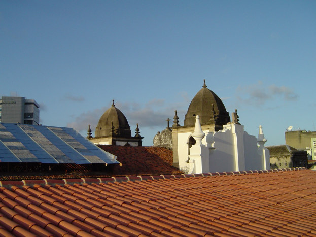 Stadtpanorama mit Gebäuden im Vordergrund, einem blauen Himmel im Hintergrund und Solarpanels auf einem Dach, die den Einsatz erneuerbarer Energien anzeigen.