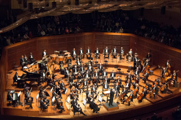 Eine Gruppe von Menschen, die auf einer Bühne sitzen und Musikinstrumente spielen, mit Buchständern davor und einem Publikum oben im Bild.