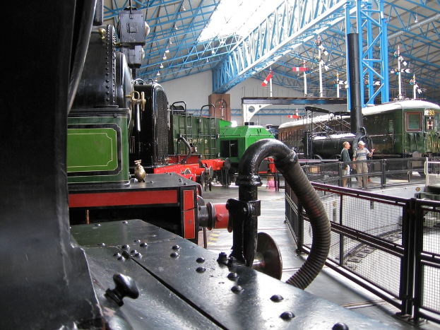 Mehrere Lokomotiven sind auf einem Abstellgleis mit zahlreichen Eisenstangen darüber geparkt, ein Zaun auf der rechten Seite, ein Zug auf dem Bahngeleise neben dem Zaun und zwei Personen, die in der Mitte neben dem Zaun stehen.