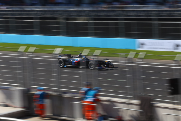 Ein fokussiertes Formel-Auto, das auf einer Rennstrecke fährt, mit verwischten Zuschauern und grasbewachsenem Hintergrund, mit Texttafeln im Hintergrund.