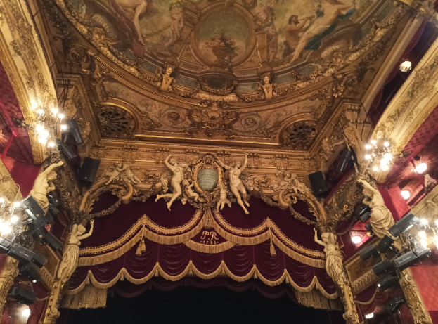Theaterinnenraum mit einer roten Gardine, Kronleuchtern, Skulpturen, Lichtern und einer Deckenmalerei, die eine Bühne und einige Menschen unten zeigt.