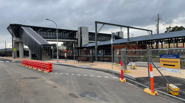 Ein Bahnhofsgebäude mit einer Brücke im Hintergrund, Straßenlaternen, Strommasten, Stromkabel, Bäume, Gebäude, Fahrzeuge auf der Straße, Absperrpoller, Schilder, Bänke und ein bewölkter Himmel.