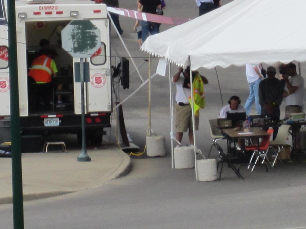 Ein Krankenwagen steht links von einer Stra√üe, w√§hrend auf der rechten Seite ein Zelt eine Gruppe von Menschen sch√ľtzt, mit einem Tisch und einem Stuhl in der N√§he.