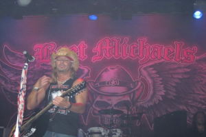 Eine Musikkonzertszene mit Bühnenbeleuchtung oben, einem Banner im Hintergrund, einer sitzenden Person in der rechten unteren Ecke, einem stehenden Gitarristen links mit einem Mikrofon davor und Gegenständen in der Mitte unten.