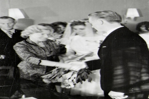 Ein Schwarz-Weiß-Foto einer Braut und eines Bräutigams, die sich die Eheringe reichen, mit der Braut in einem weißen Kleid und Schleier, die einen Blumenstrauß hält, und dem Bräutigam im Anzug und Krawatte, mit Wandlampen im Hintergrund.