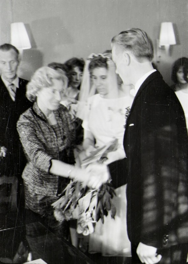 Ein Schwarz-Weiß-Foto einer Braut und eines Bräutigams, die sich die Eheringe reichen, mit der Braut in einem weißen Kleid und Schleier, die einen Blumenstrauß hält, und dem Bräutigam im Anzug und Krawatte, mit Wandlampen im Hintergrund.