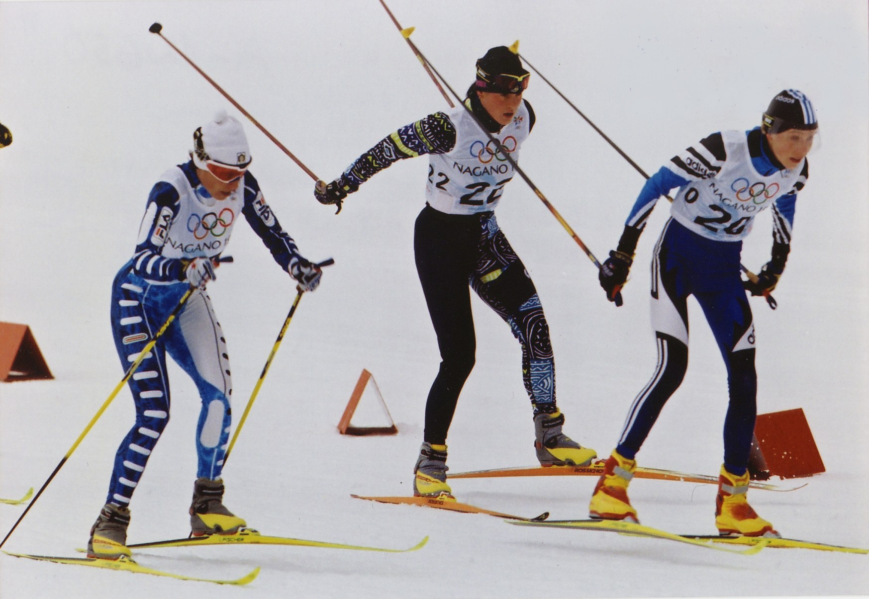 Drei Skifahrer in bunten Anzügen und Helmen rasen eine glitzernde schneebedeckte Piste hinunter, jeder hält Ski-Stöcke.