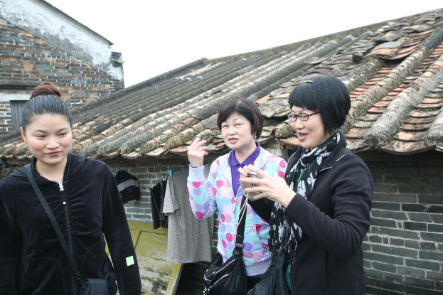 Drei Frauen stehen zusammen, wobei zwei lÃ¤cheln und eine eine Brille trÃ¤gt, vor HÃ¤usern unter einem Himmel.