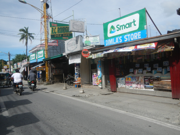 Eine belebte Straße mit Motorrädern, Autos, Gebäuden, Schaufenstern, Schildern, Strommasten, Kabeln, Straßenlaternen, Bäumen und einem bewölkten Himmel.